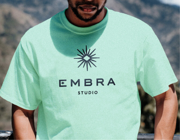 Embra logo on a mint green tee worn by a young black man in a mountainous setting.