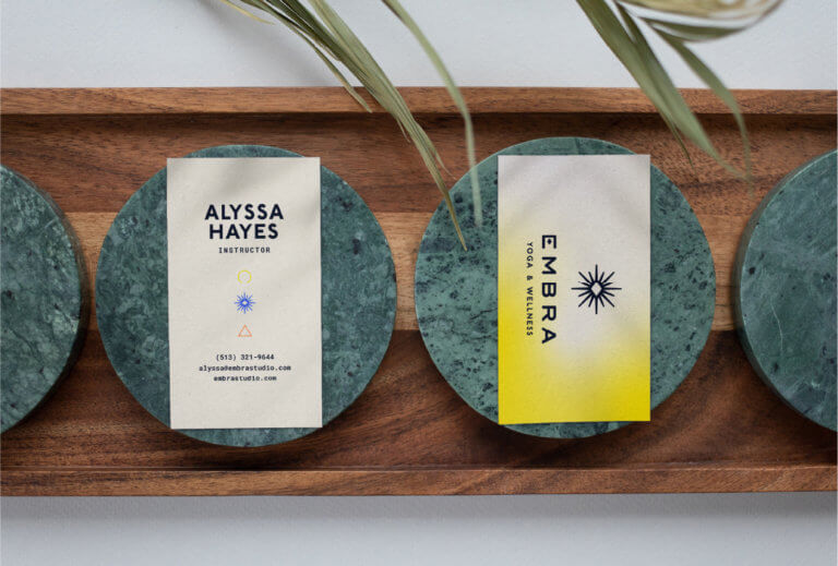 Business cards branded with the Embra logo backed by a yellow gradient. The business cards sit on green marble coasters placed along a wooden tray.