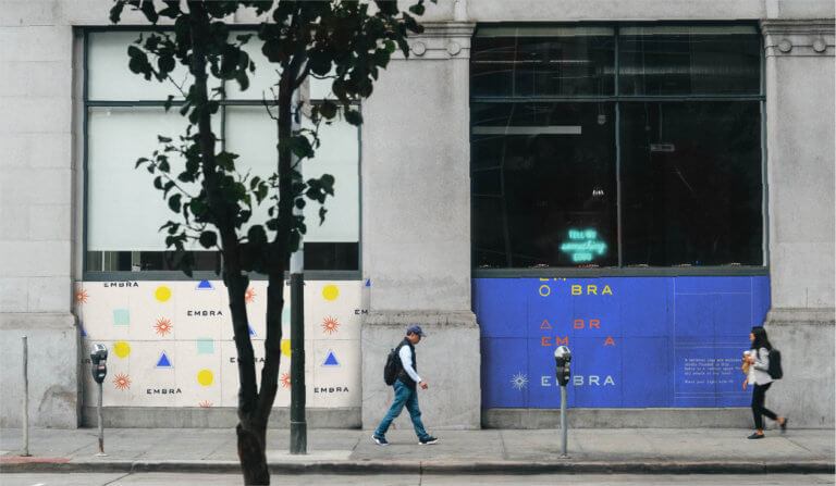Vibrant Embra-branded wall billboards brighten up and urban street with passersby and trees in the foreground