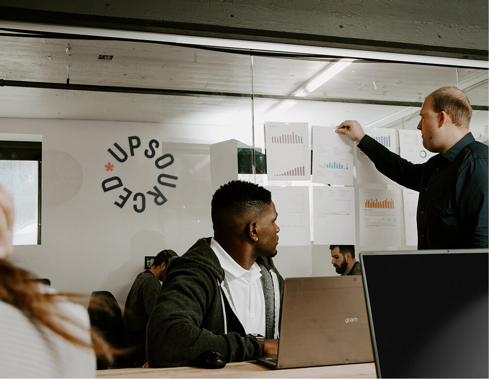 A team meeting takes place in a room surrounded with glass walls with the Upsourced logo on one of the walls.