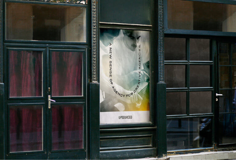 An Upsource branded wheatpaste poster is nestled between to storefront windows in an urban environment.